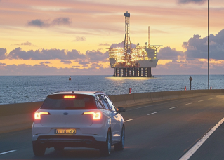 Een witte auto op aardgas rijdt langs een kustweg bij zonsondergang. Op de achtergrond is een platform te zien waar aardgas wordt gewonnen.