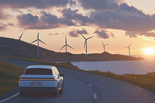 Een witte hybride auto rijdt bij zonsondergang over een kustweg. Aan de horizon zijn zes windturbines te zien.