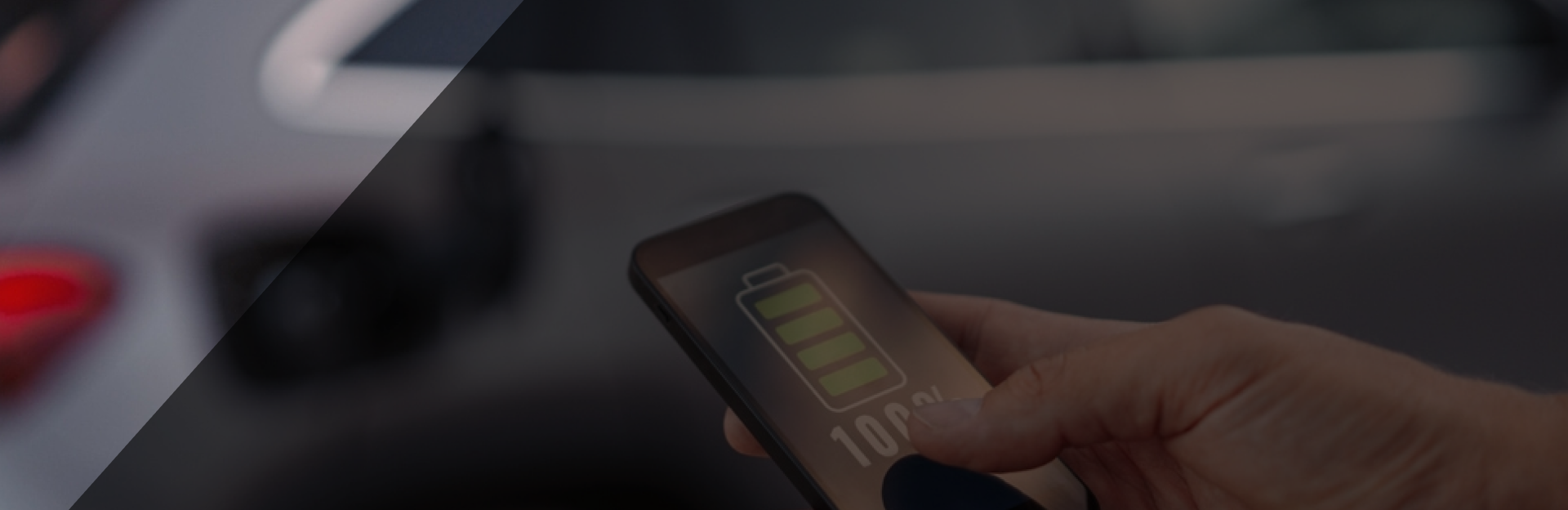 Hand met een telefoon waarop een 100% opgeladen batterij te zien is. Op de achtergrond is een elektrisch voertuig te zien.